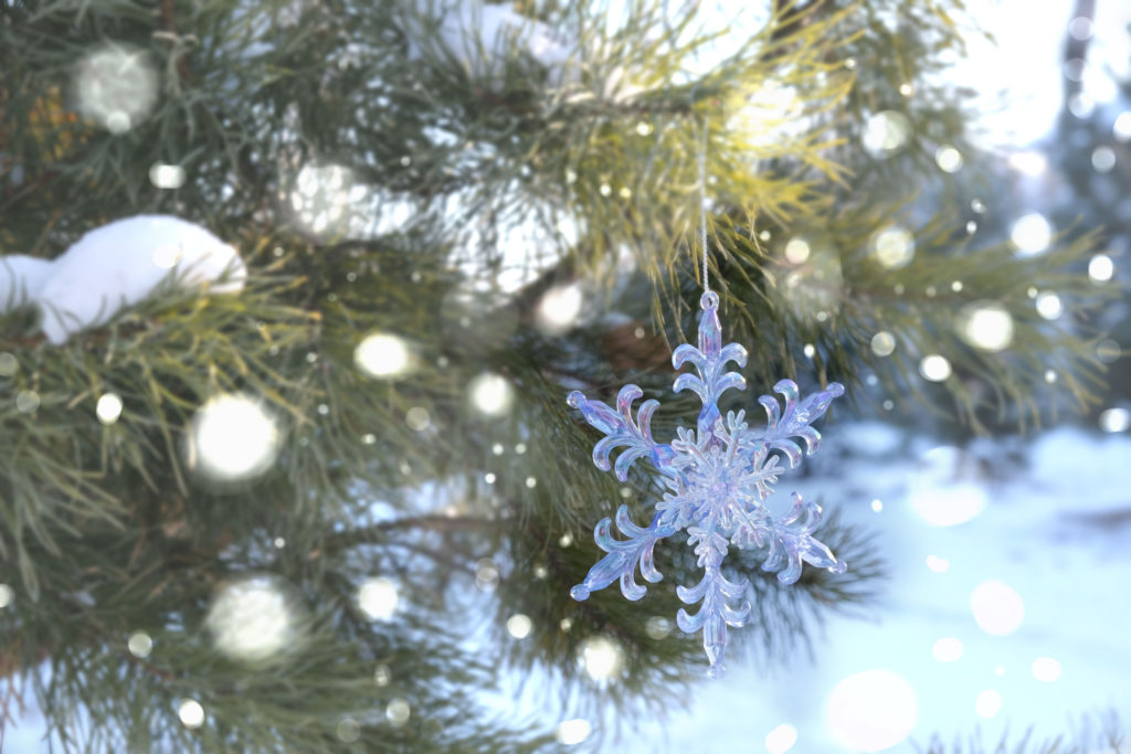 A Christmas tree outside covered in show with a snowflake ornament hanging from a branch