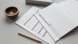 Finance tracker workbook open a table next to a bowl of paper clips and a gold pen