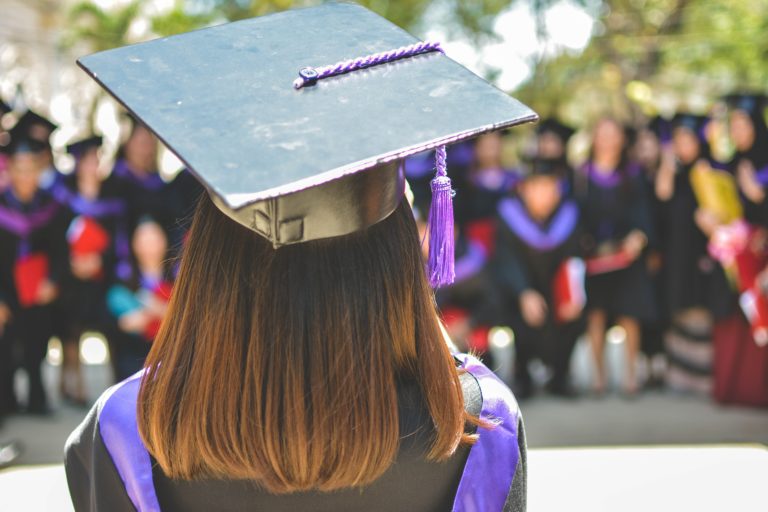 The back of a womans head who is wearing a graduation gap. Her fellow graduates are our of focus in the background.