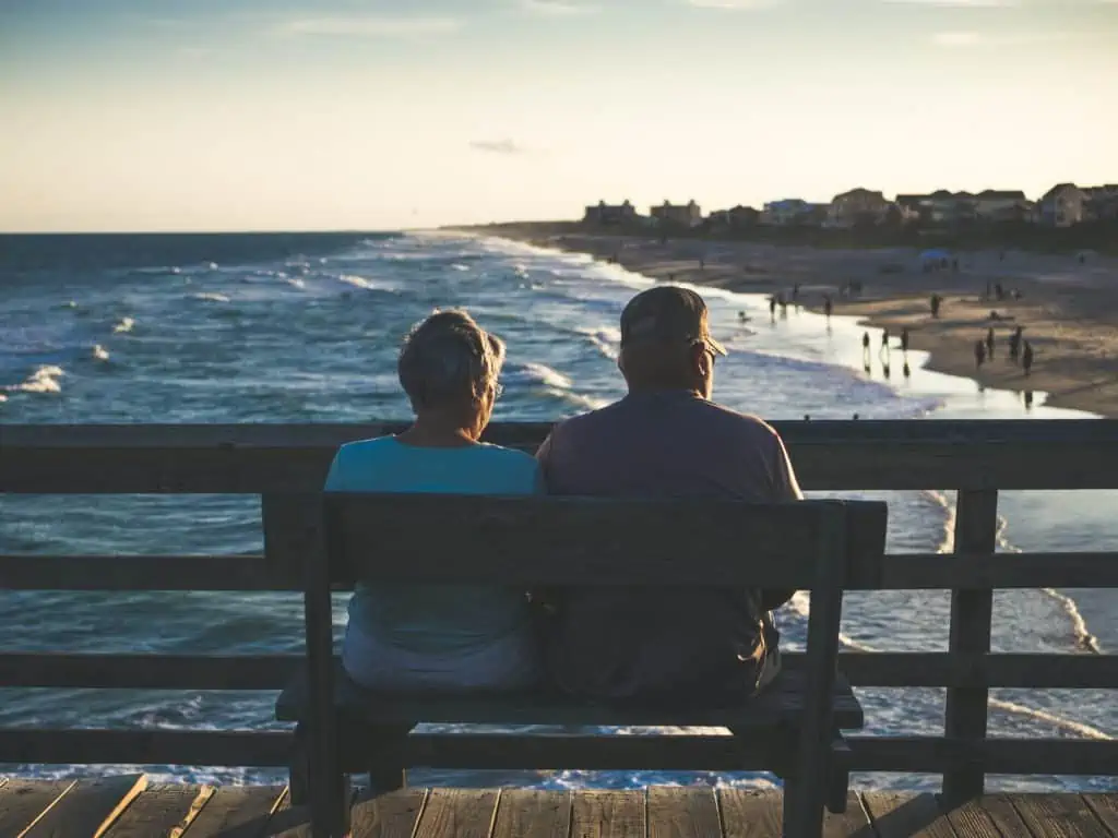 An older couple sitting on a bench on the boardwalk overlooking the beach